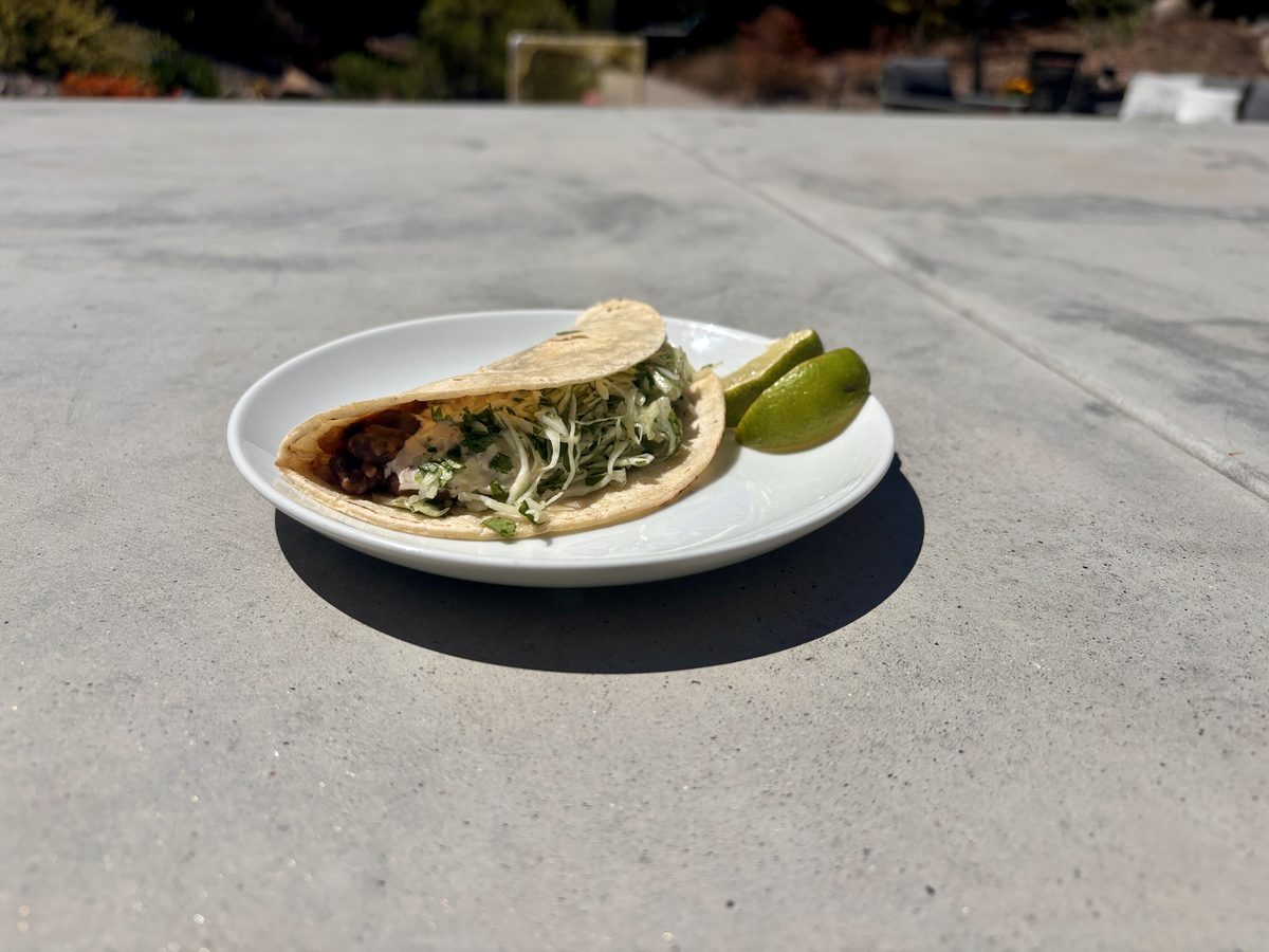 One folded soft corn tortilla taco filled with a dark bean mixture and shredded lettuce, with a lime wedge beside it on a white pl