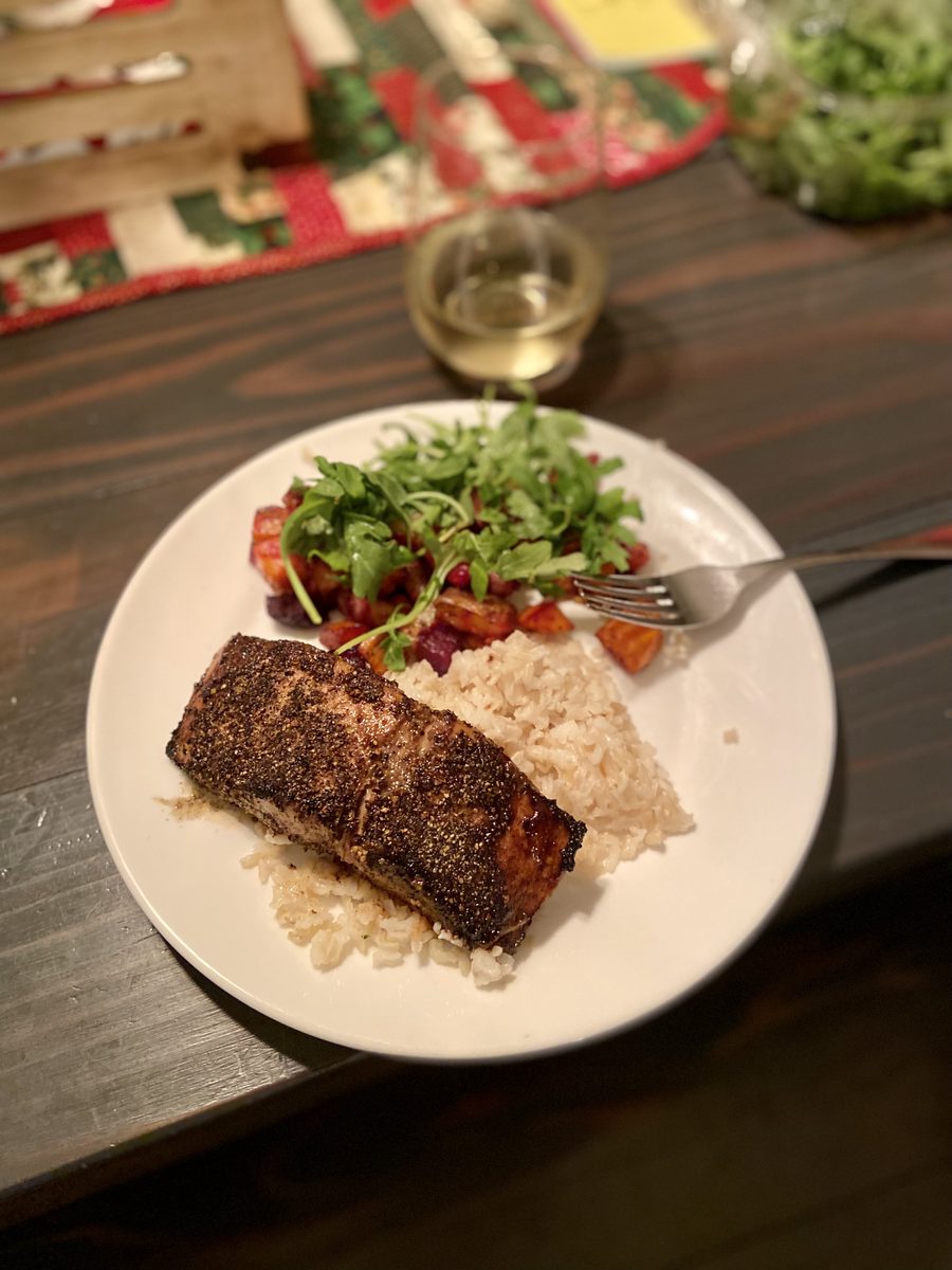 A blackened salmon fillet with brown rice and an arugula-and-tomato salad on a white plate, fork on the rim, on a dark wood table 