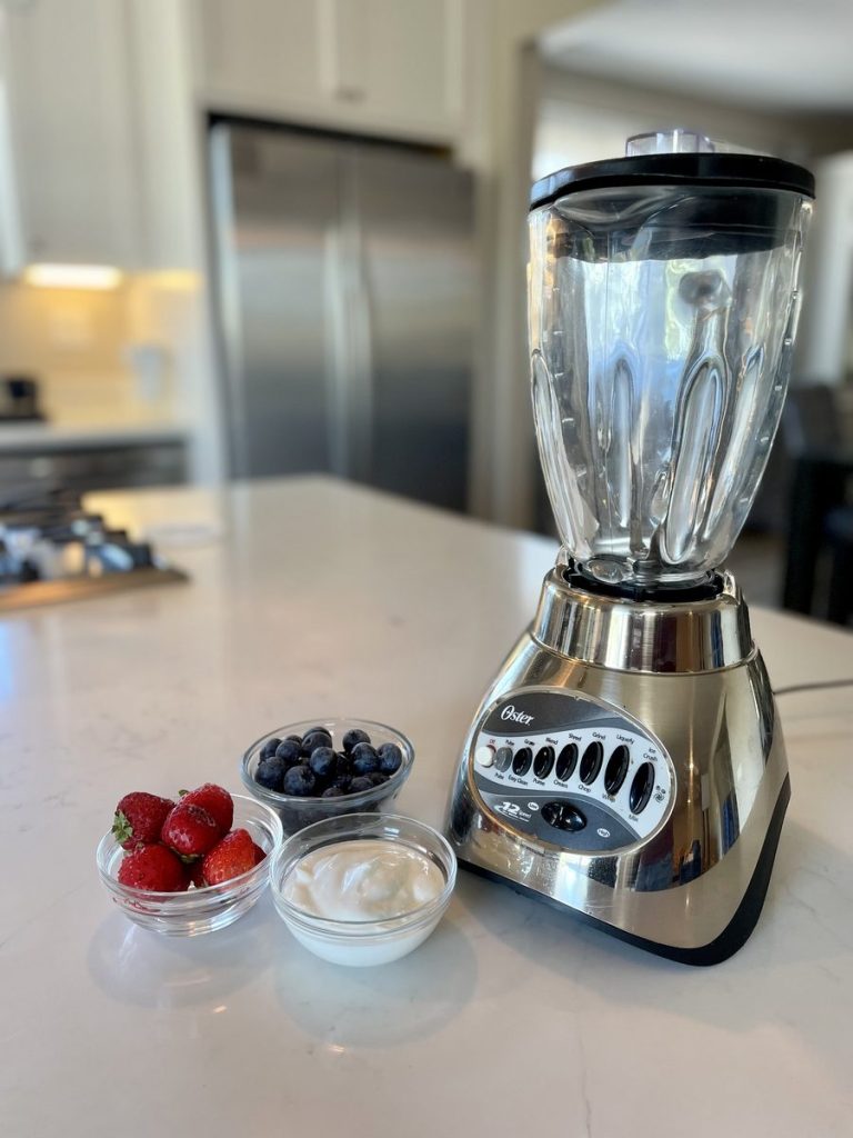 An empty stainless countertop blender on a white kitchen island with small glass bowls of strawberries, blueberries and yogurt beside it; stainless fridge and c