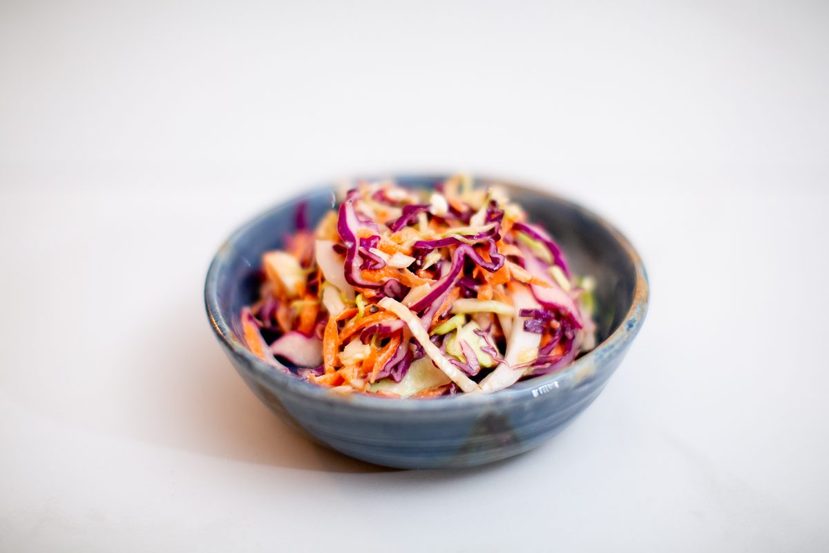 A speckled stoneware bowl of shredded purple and green cabbage slaw with carrot, against a plain white background.