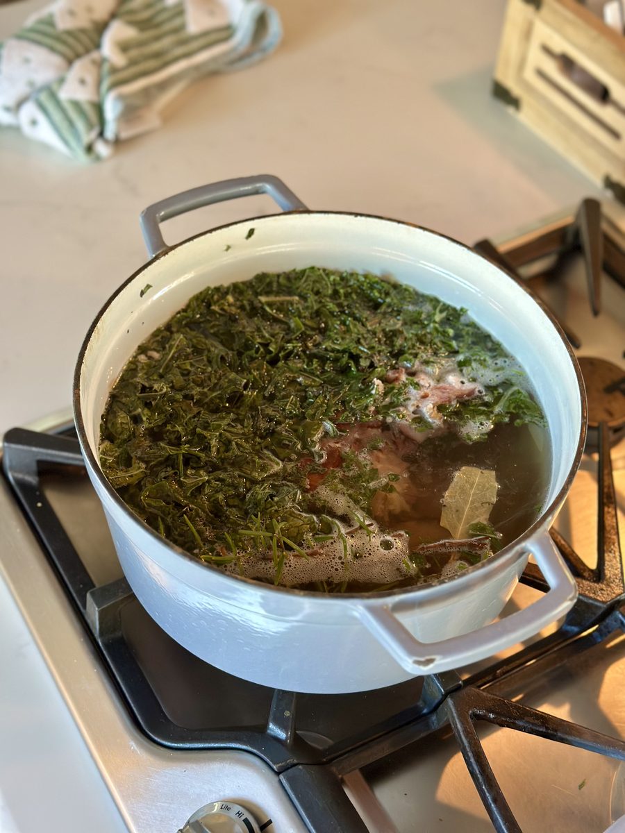 A white enamelled dutch oven on a gas range holding simmering broth thick with chopped green herbs, pale poultry pieces and a bay