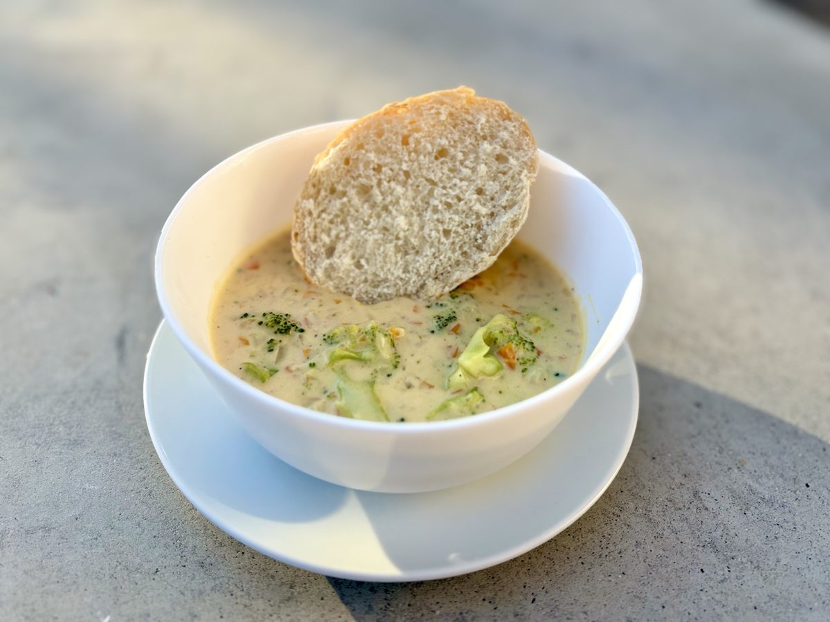 A white bowl on a saucer holding a thick pale green-flecked creamy soup with broccoli florets, a torn round of crusty bread balanc