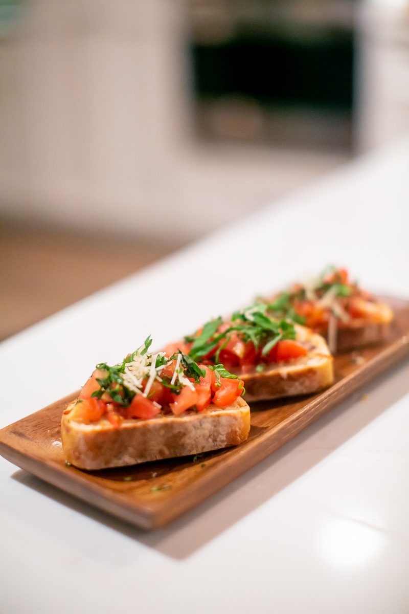 Three bruschetta toasts topped with diced tomato, arugula and shaved parmesan on a narrow wooden serving board on a white counter,