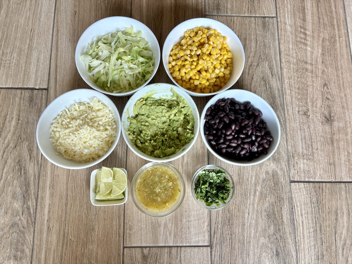 Overhead cluster of white bowls holding shredded cabbage, corn, cotija cheese and guacamole on grey wood