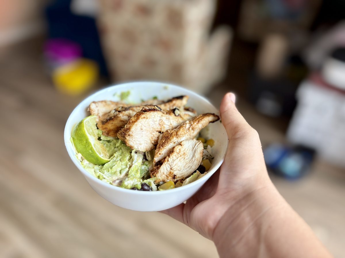 A hand holding a prepped burrito bowl with grilled chicken, black beans, corn, guacamole and lime.