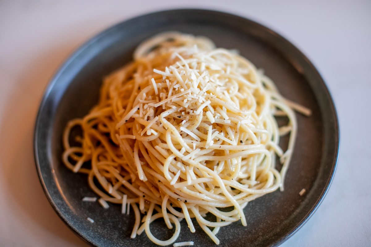 A twirled mound of buttered spaghetti topped with grated parmesan on a dark speckled ceramic plate against a plain pale background