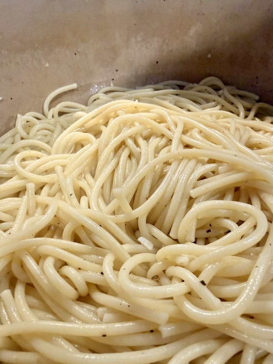 Extreme overhead close-up filling the frame with glossy buttered spaghetti flecked with black pepper, in a pale pot or bowl with n