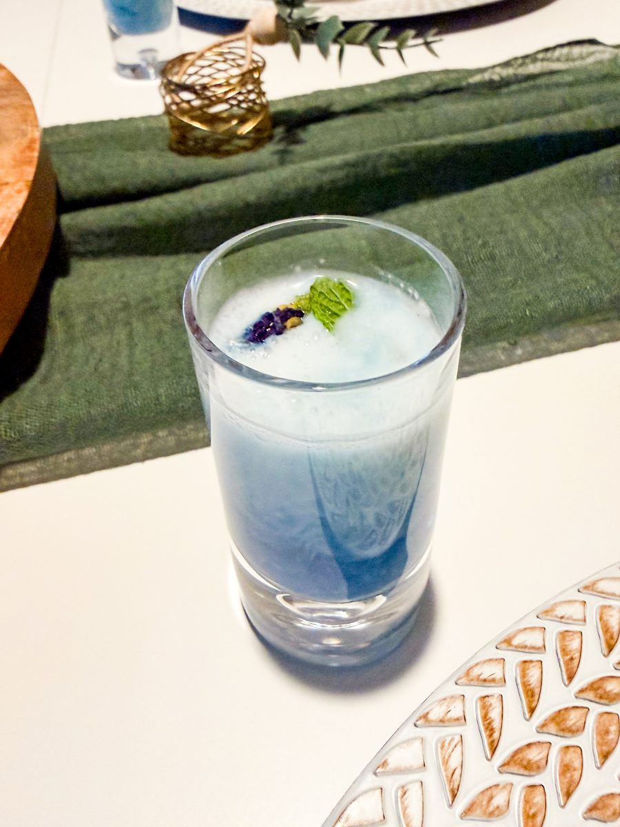 A tall glass of layered blue butterfly pea tea with coconut milk, garnished with a mint sprig and a purple flower, on a white tabl