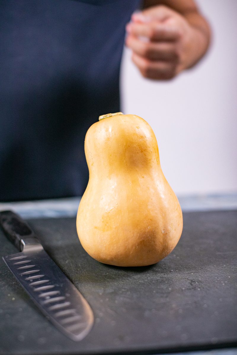 A whole butternut squash standing upright on a dark stone board next to a chef's knife, with a person's navy-aproned torso out of 