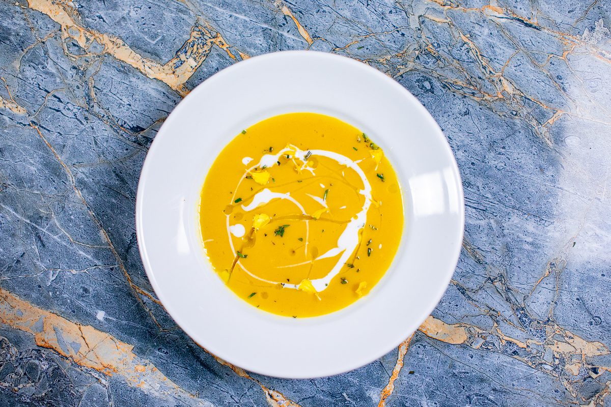 An overhead shot of a white bowl of smooth orange soup swirled with cream and flecked with herbs, on blue marble.