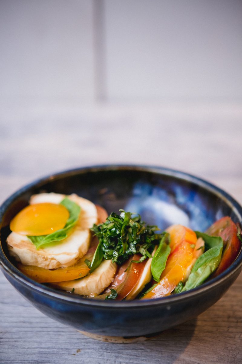 A dark blue bowl of caprese-style salad with heirloom tomato slices, fresh mozzarella, basil and herb dressing topped with a fried