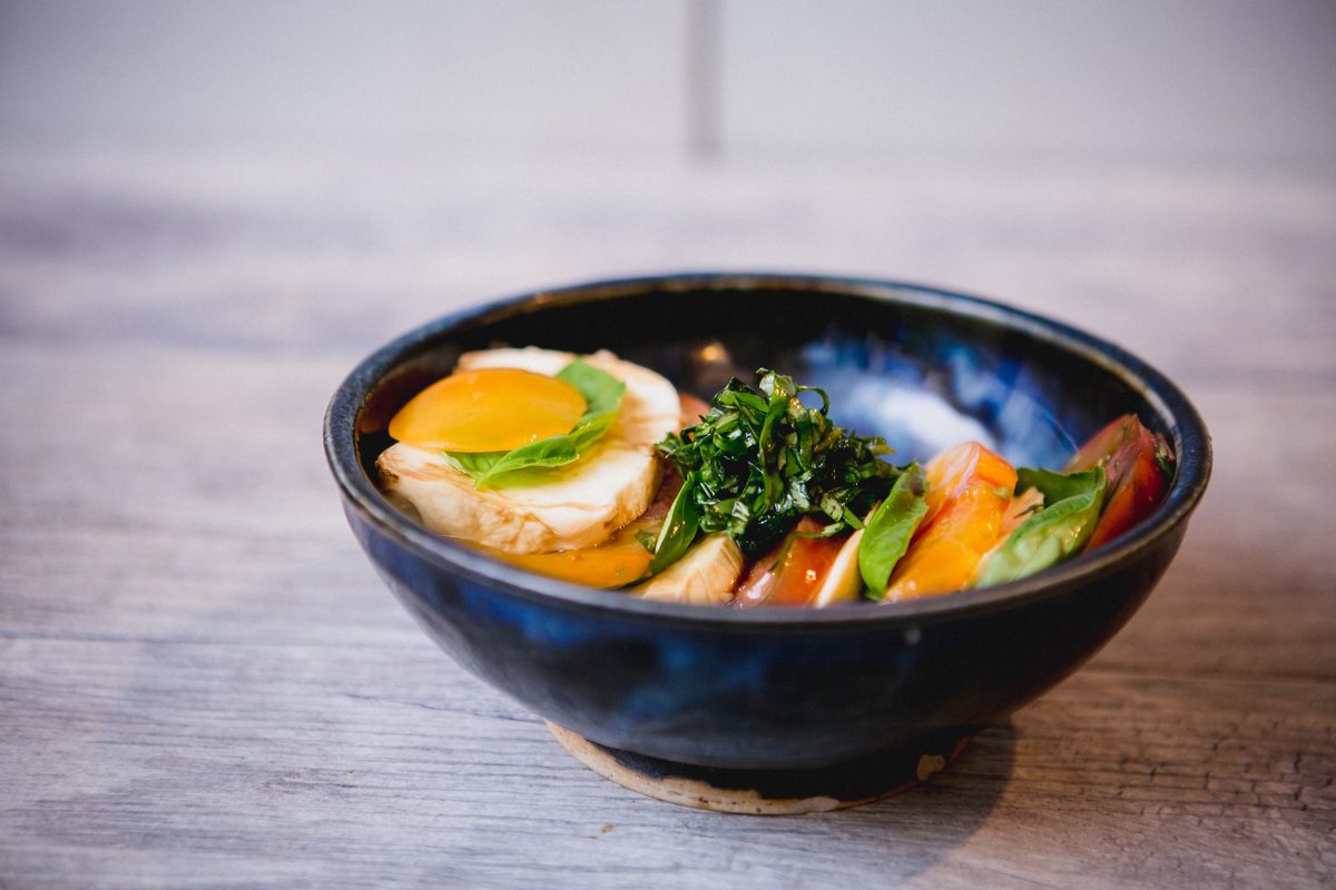 Side view of a navy bowl holding tomato, mozzarella and basil salad crowned with a runny fried egg, on gray wood planks.