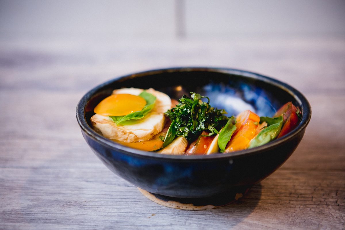 A dark ceramic bowl of layered tomato, mozzarella, basil and greens with a fried egg on top, shot at table level on wood.