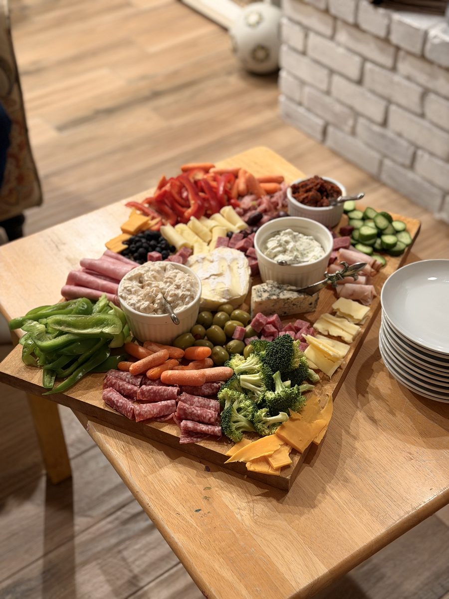 A large wooden charcuterie board on a small wood table loaded with rolled salami and folded deli meats, sliced cheddar and other c