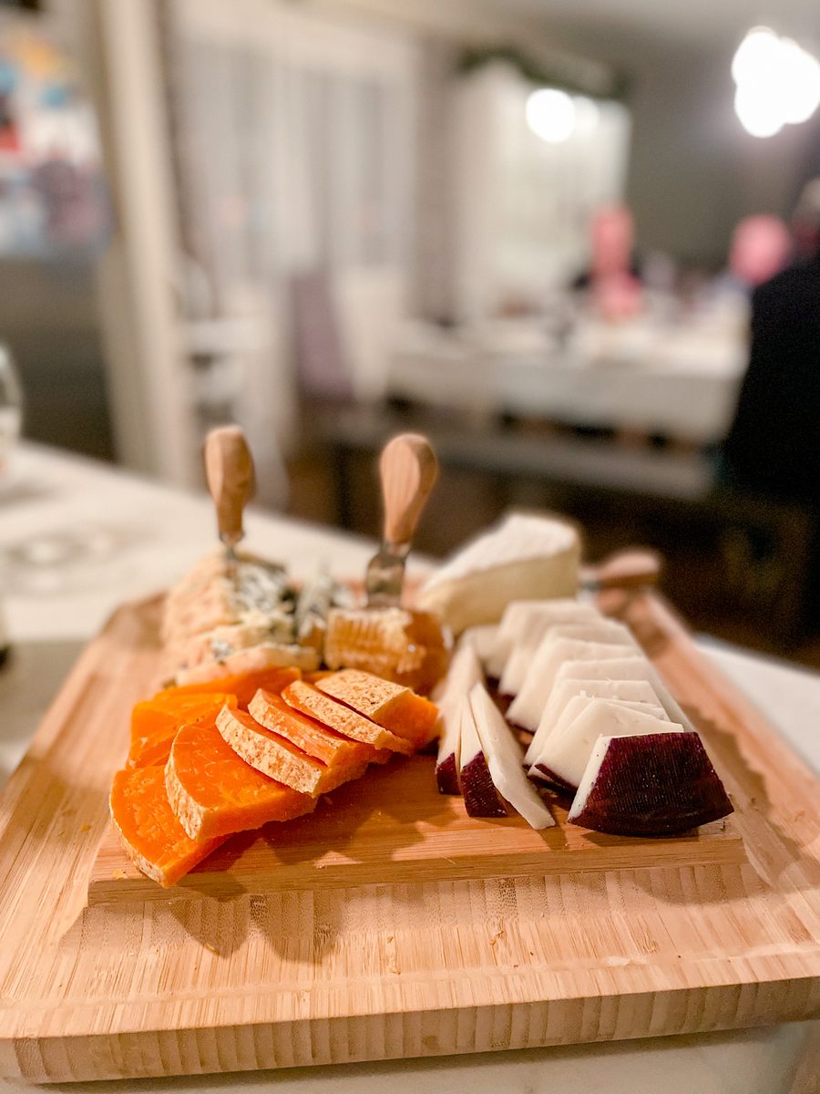 A bamboo cutting board holding sliced orange cheddar-style cheese, a blue-veined wedge, brie and a dark red-rind cheese with two s