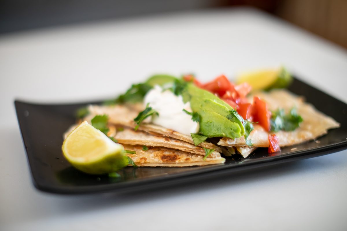 Quesadilla wedges fanned on a black rectangular plate, topped with sour cream, sliced avocado, diced tomato pico de gallo and cila
