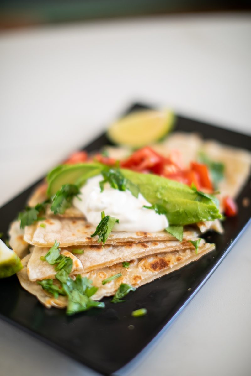 A tighter angled view of the same cheese quesadilla wedges on a black rectangular plate with sour cream, avocado slices, diced tom