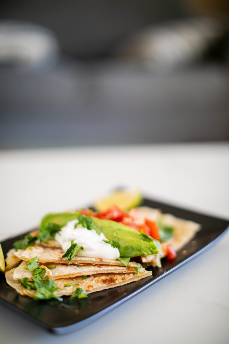 Cheese quesadilla wedges on a black rectangular plate topped with sour cream, avocado, diced tomato and cilantro, shot low across 