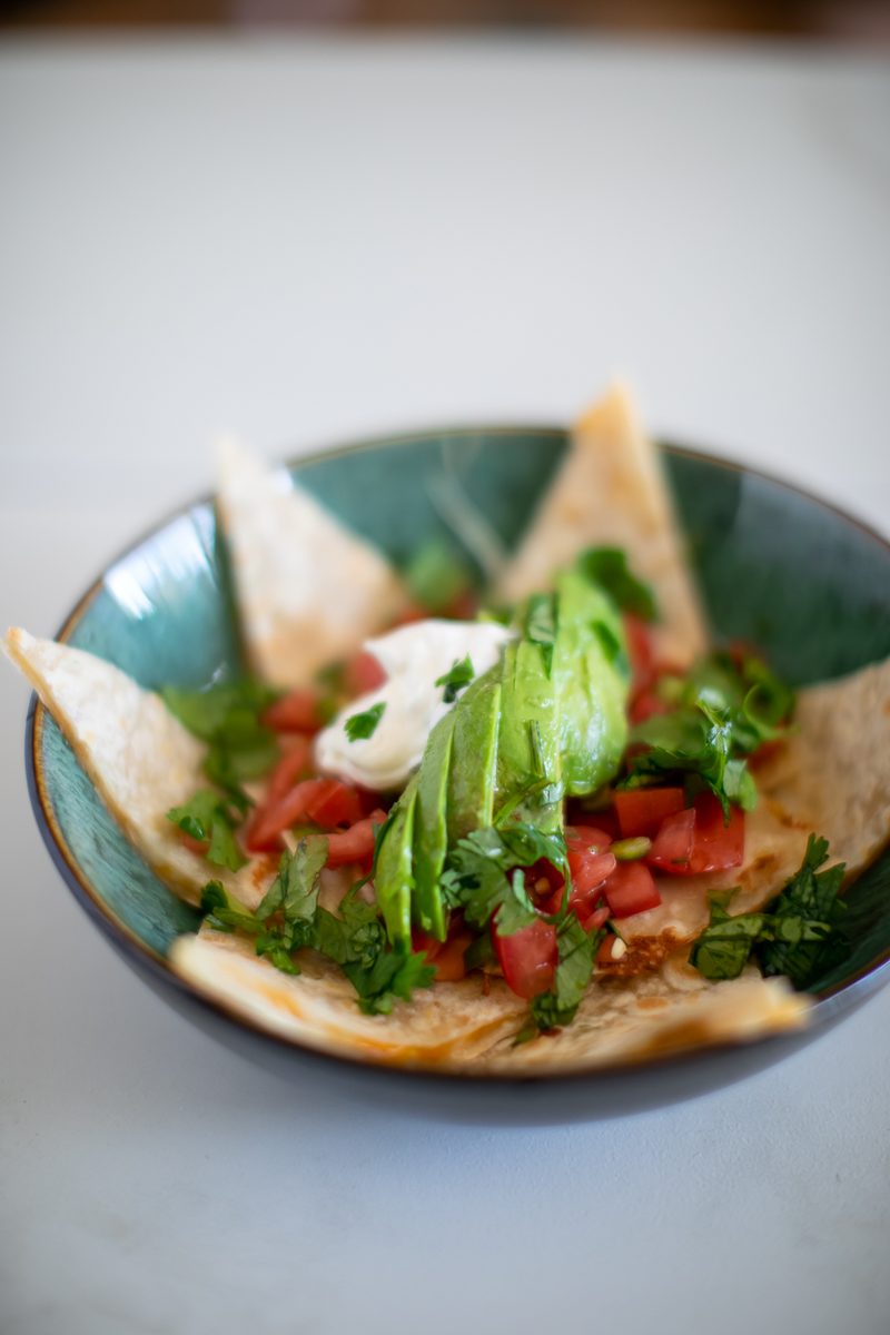 A teal ceramic bowl of quesadilla wedges topped with sour cream, sliced avocado, diced tomato and plenty of cilantro, on a pale gr