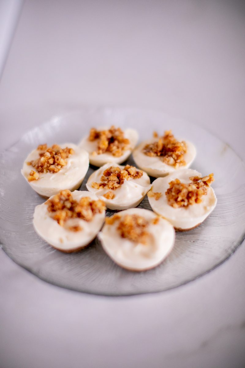 Seven mini cheesecakes with pale cream tops and a chopped nut streusel topping arranged on a clear glass plate against a pale lave