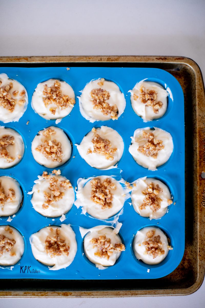 A blue silicone muffin pan on a baking sheet holding a dozen small white baked custards topped with crumbly nut topping, still in 