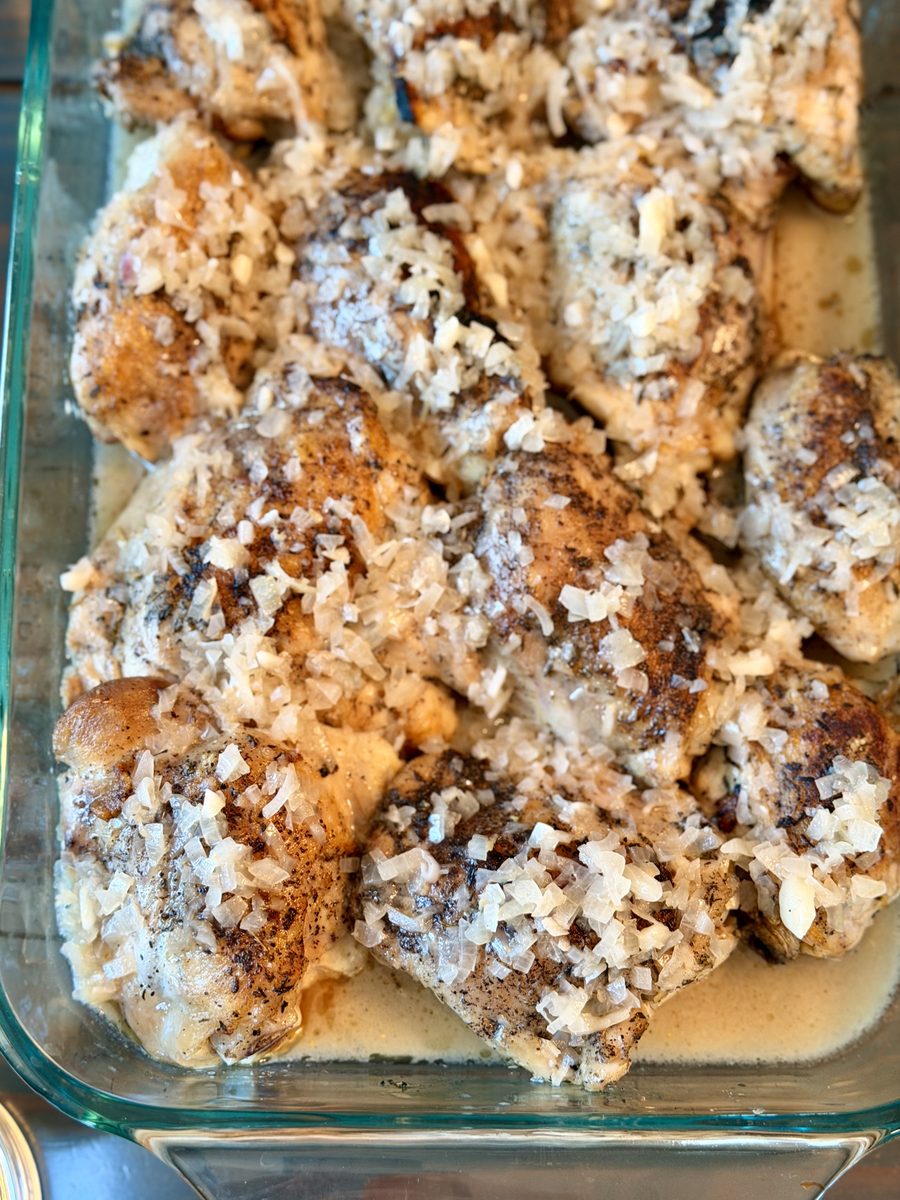 Browned bone-in chicken pieces baked in a clear glass casserole dish, covered in melted cheese and herb crumbs with pan juices poo