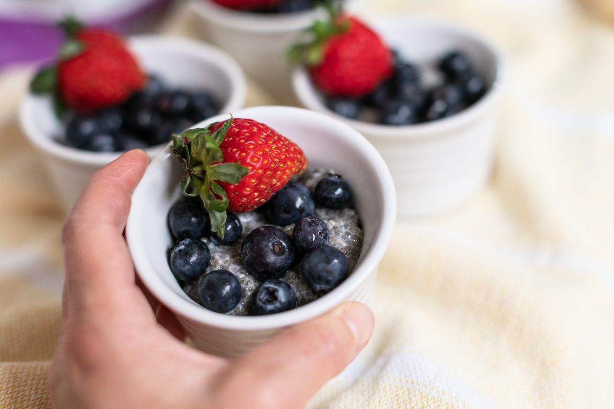 A hand holds a small white ramekin of chia pudding topped with blueberries and a strawberry, more ramekins and a cream cloth behin