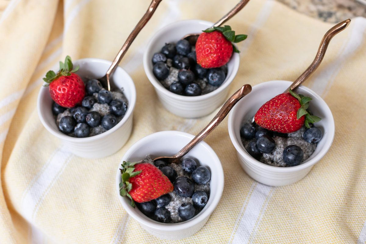Three white ramekins of chia pudding topped with blueberries and strawberries with copper spoons, arranged on a pale yellow cloth.