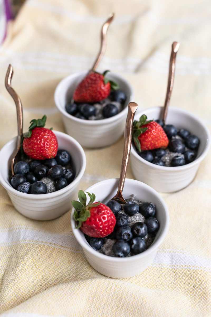 Three ramekins of chia pudding heaped with blueberries and strawberries, copper spoons standing upright, on a cream linen cloth.