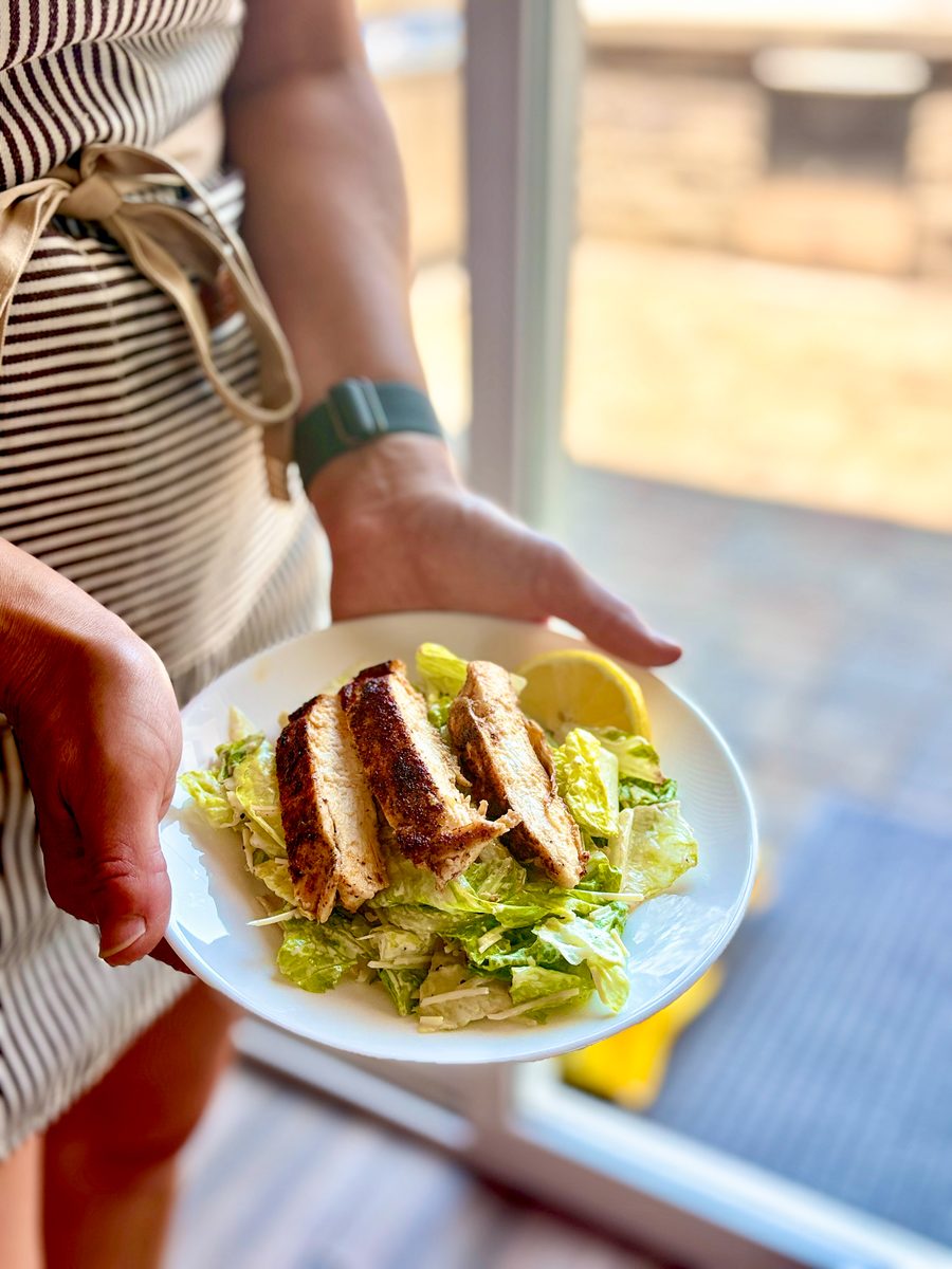 Hands hold a white plate of caesar salad topped with sliced grilled chicken breast and a lemon wedge, backlit by a patio door.
