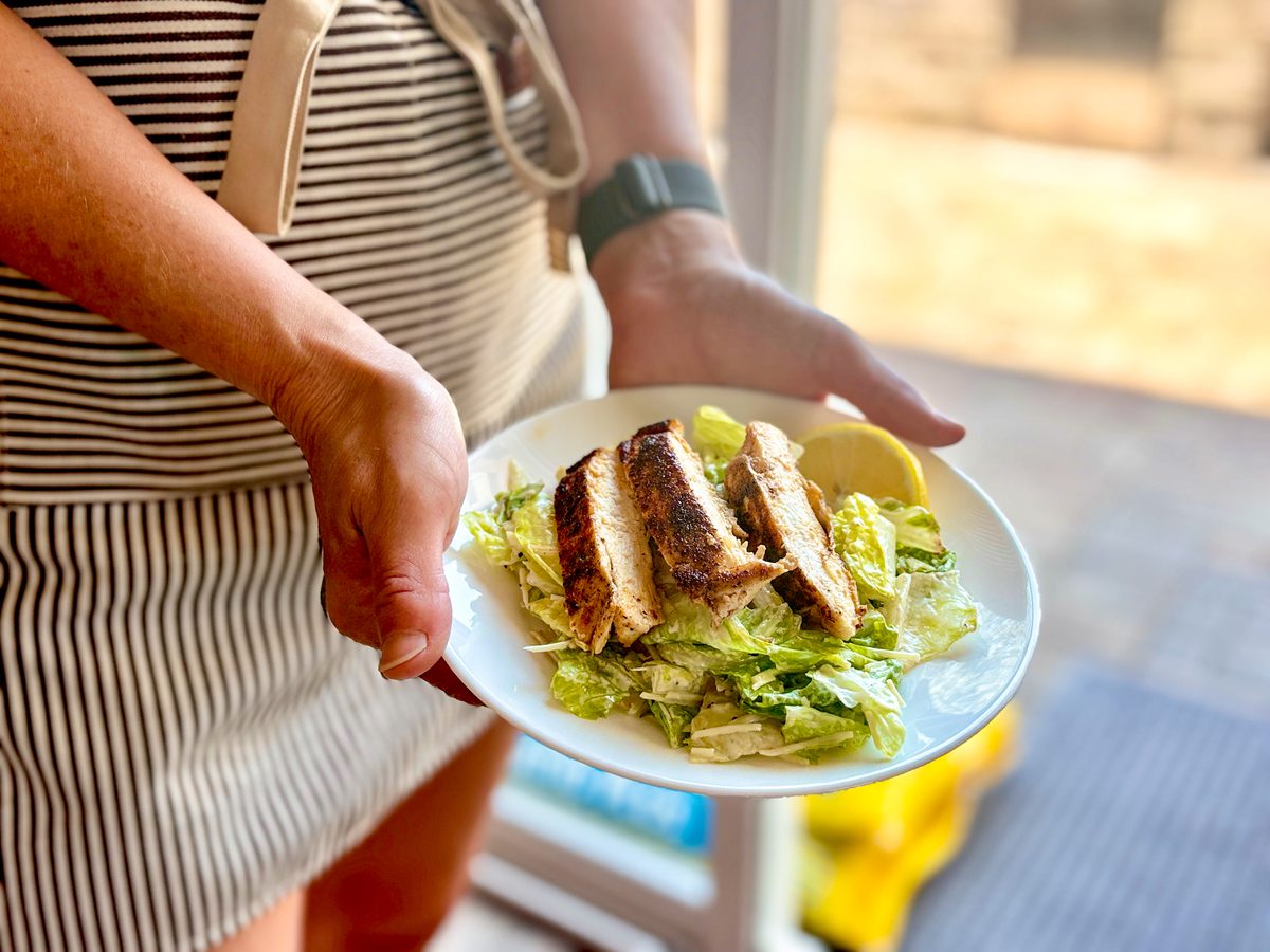 A person in a striped apron holds out a plate of caesar salad topped with sliced grilled chicken and lemon, outdoors in bright lig