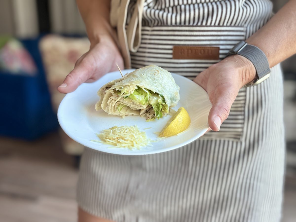 Close view of hands presenting a white plate holding a rolled caesar-style tortilla wrap filled with chopped lettuce, with a lemon