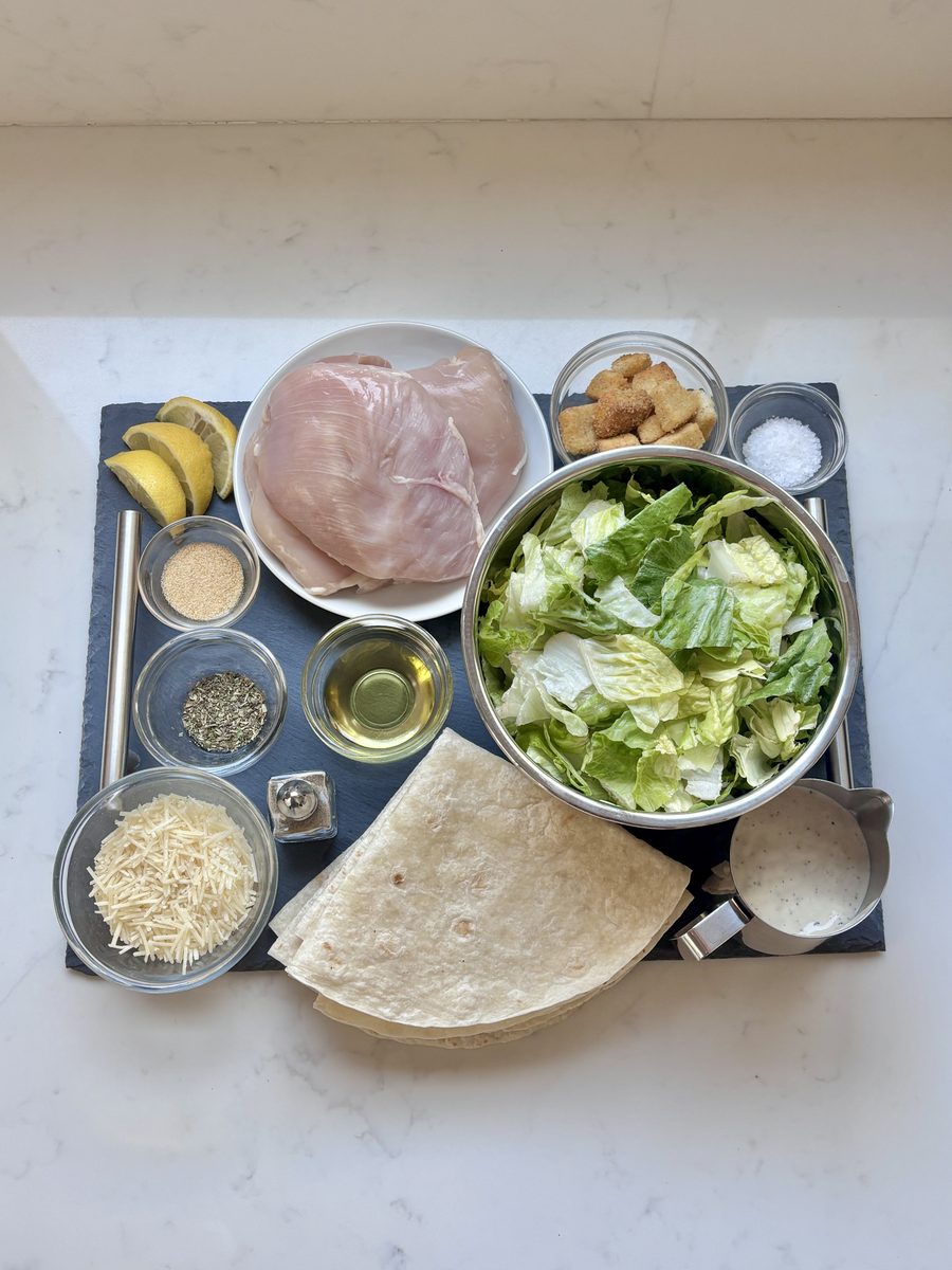 An overhead flat lay of wrap ingredients on white marble - a raw chicken breast and chopped romaine in metal bowls set in a muffin