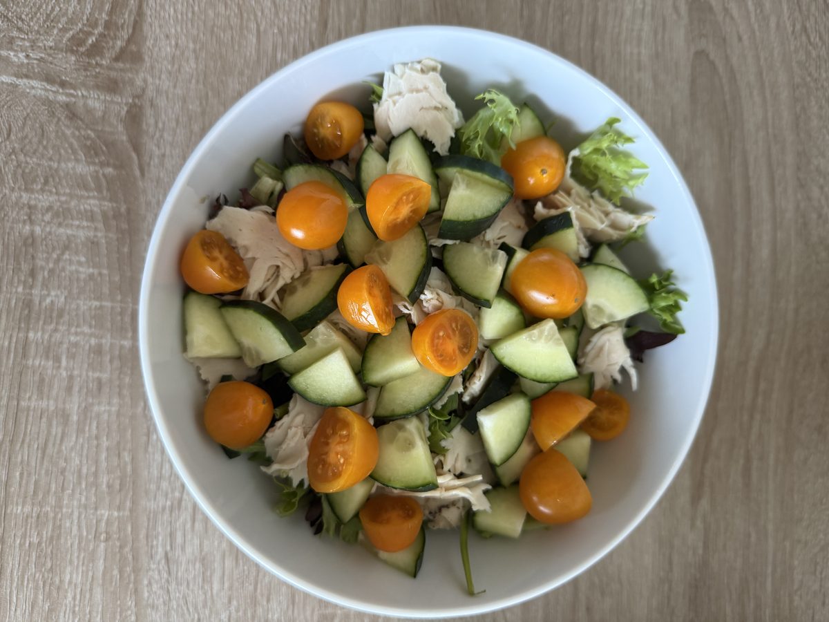 A white bowl of salad on a wood table with chunked cucumber, halved orange cherry tomatoes, mixed greens and pieces of pale shredd