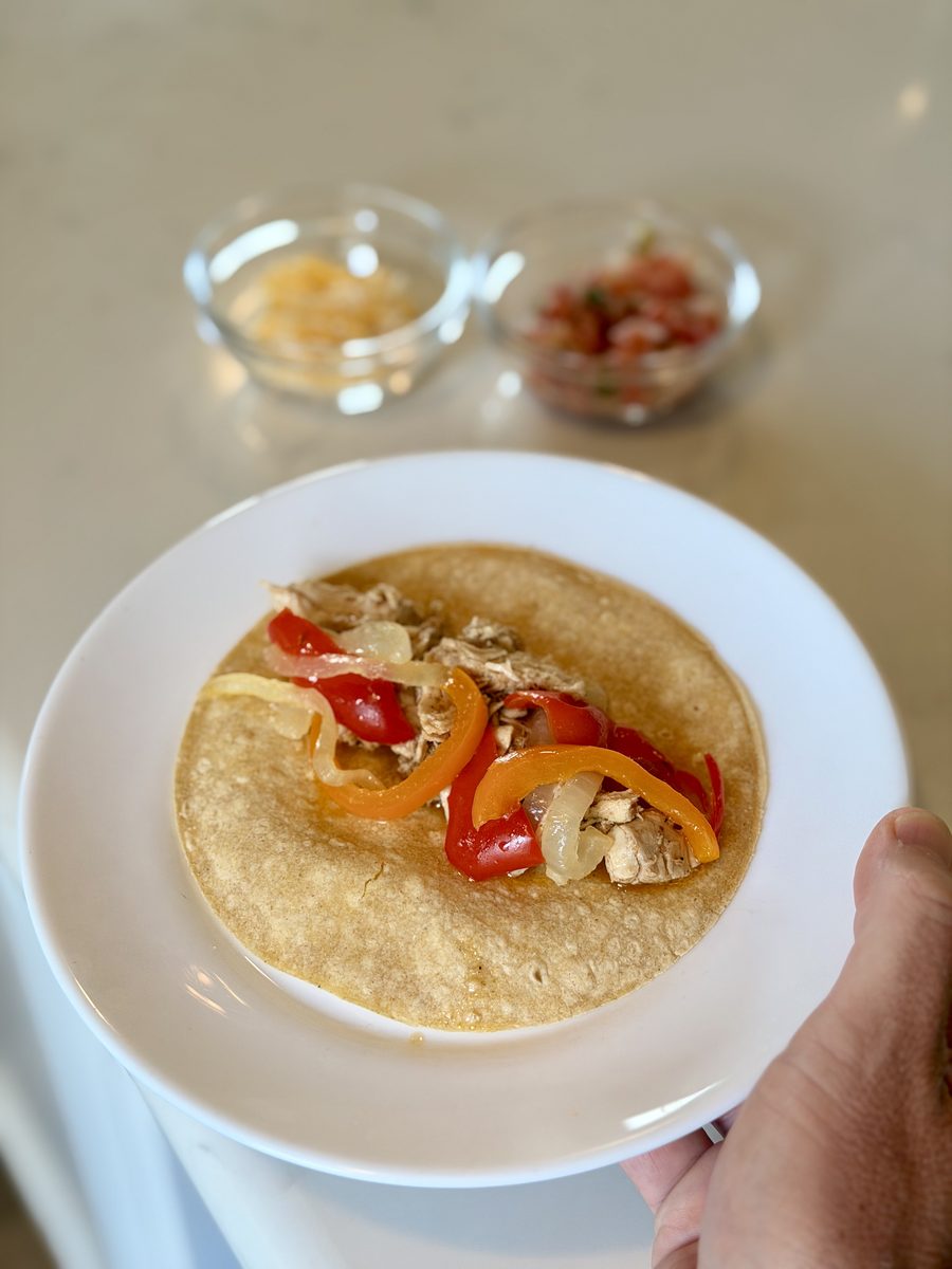 A hand holds a white plate with an open flour tortilla topped with strips of red and yellow bell pepper, onion and small pale chun