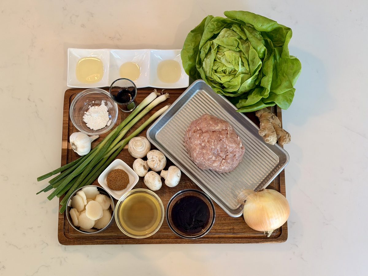 An overhead flat lay on a wooden board over white marble - a mound of raw ground chicken on a metal tray, a head of butter lettuce