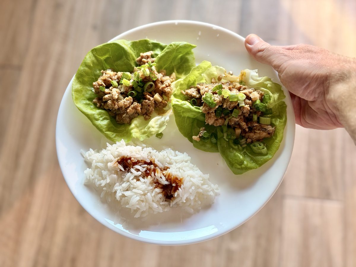 A hand holds a white plate with three butter lettuce cups filled with ground chicken and scallions plus a scoop of white rice with