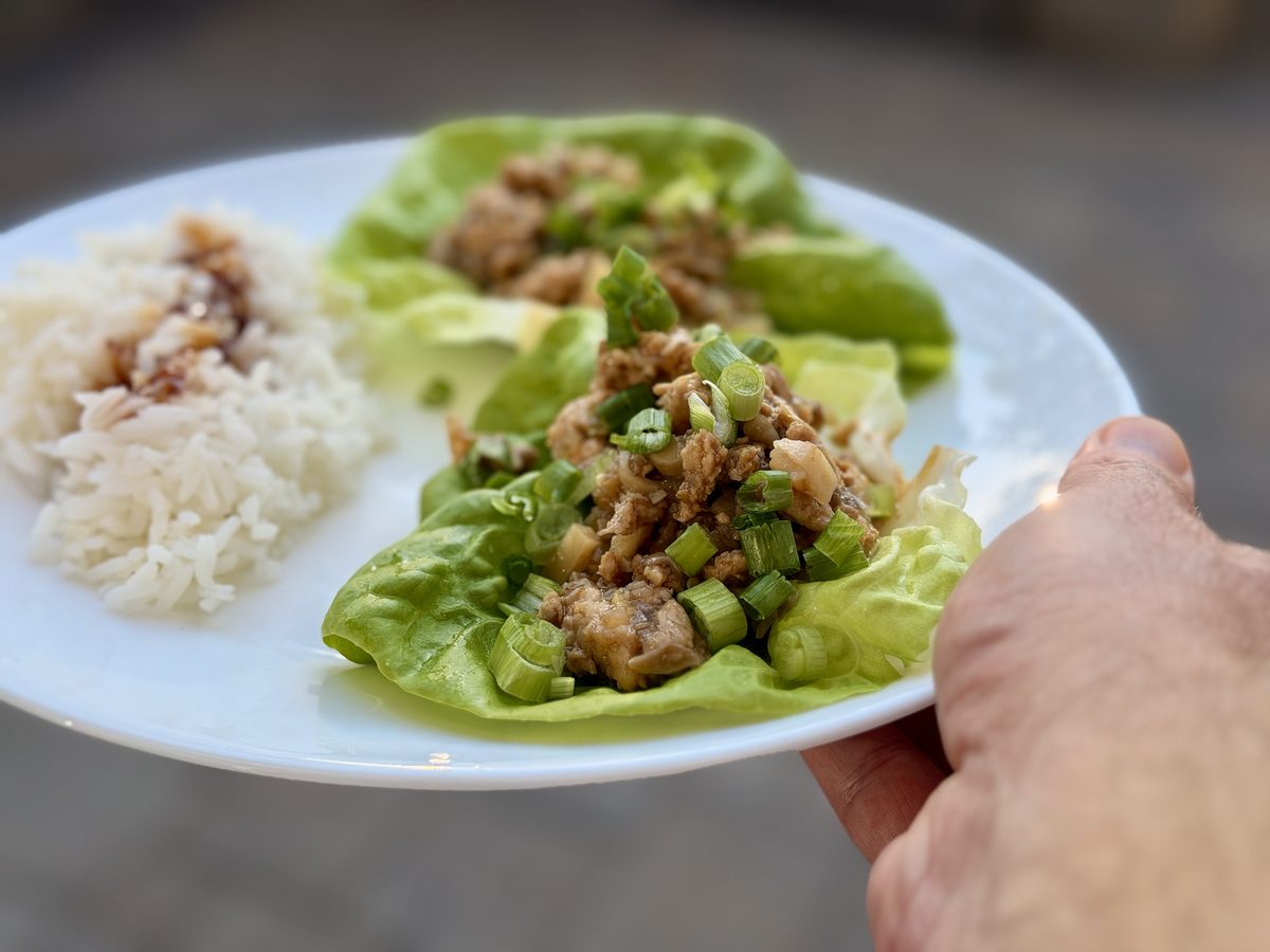 A hand holds a white plate with two chicken-filled butter lettuce cups and a scoop of white rice with drizzled sauce, outdoors.