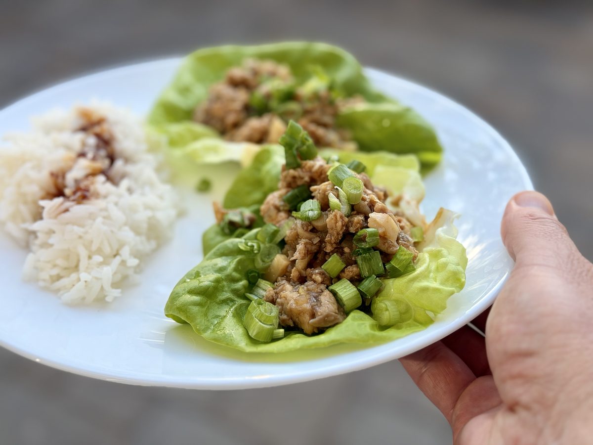 A hand holds a white plate with two ground chicken lettuce cups and a serving of white rice with sauce, outdoors on a wood deck.