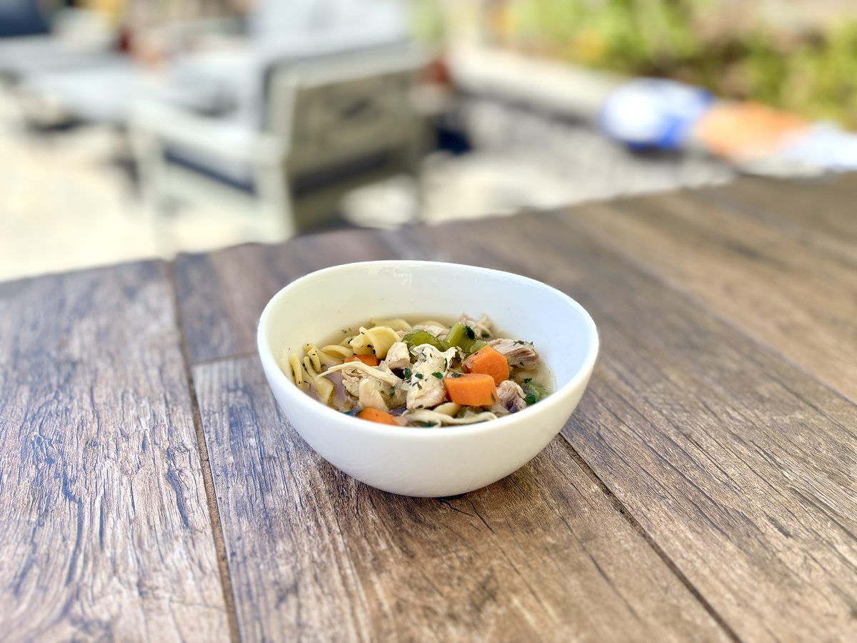 A white bowl on a weathered wood table filled with clear broth, egg noodles, sliced carrots, celery and chunks of white chicken.