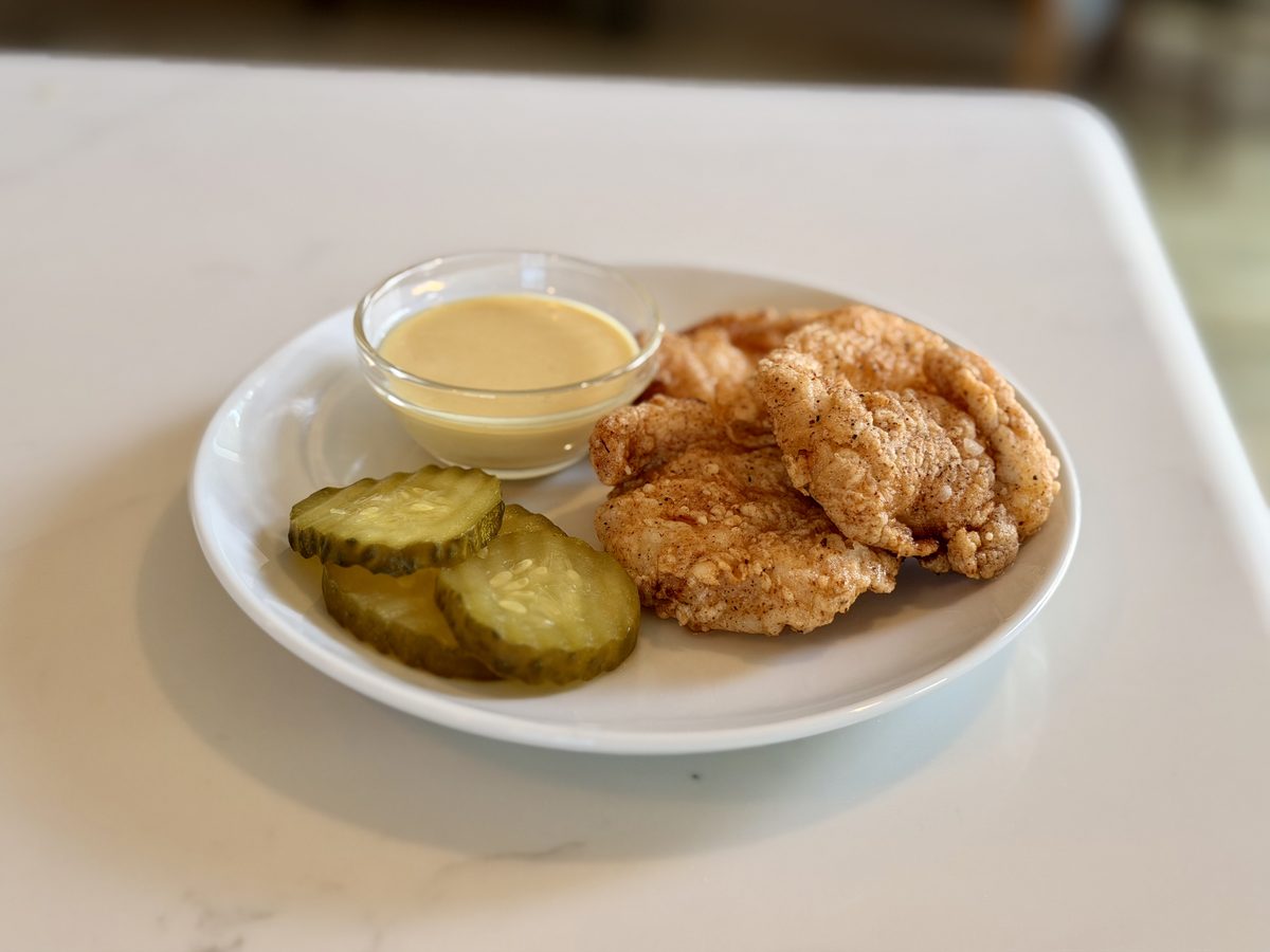 A white plate with several craggy fried chicken pieces, three pickle slices and a small dish of pale dipping sauce.