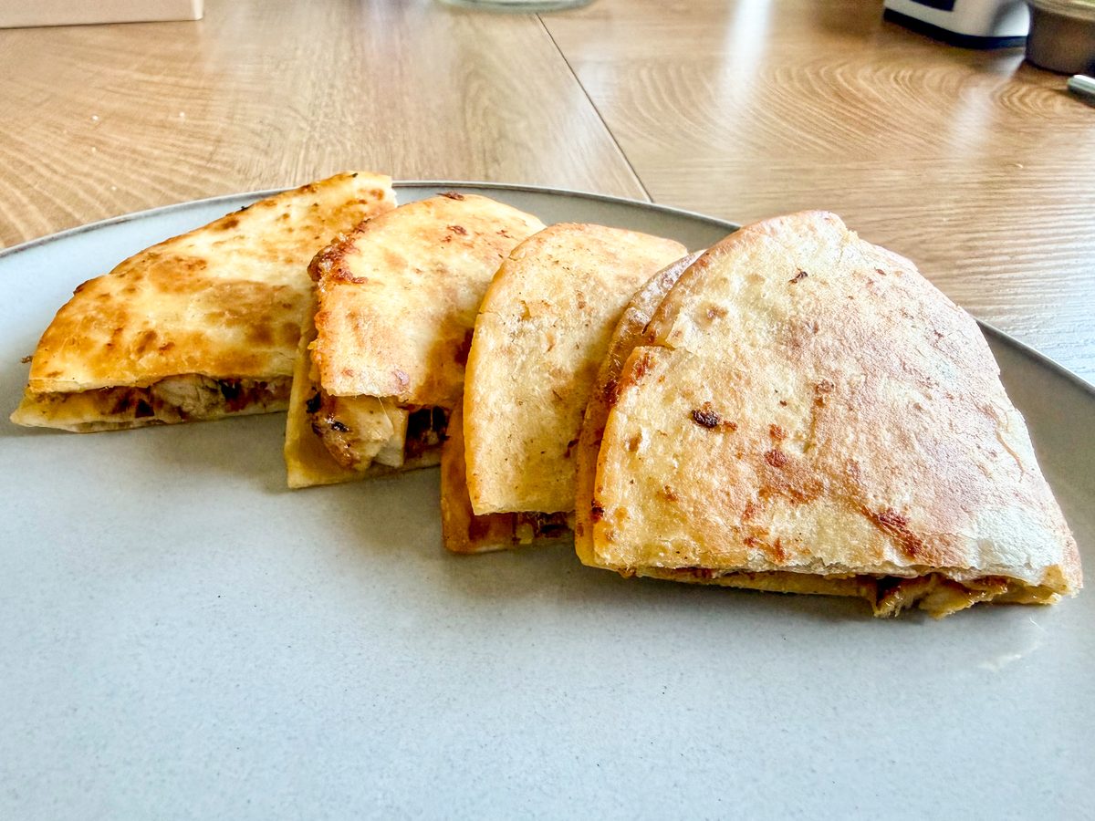 Four browned quesadilla wedges fanned on a grey plate on a light wood table, cut edges showing shredded chicken and melted cheese