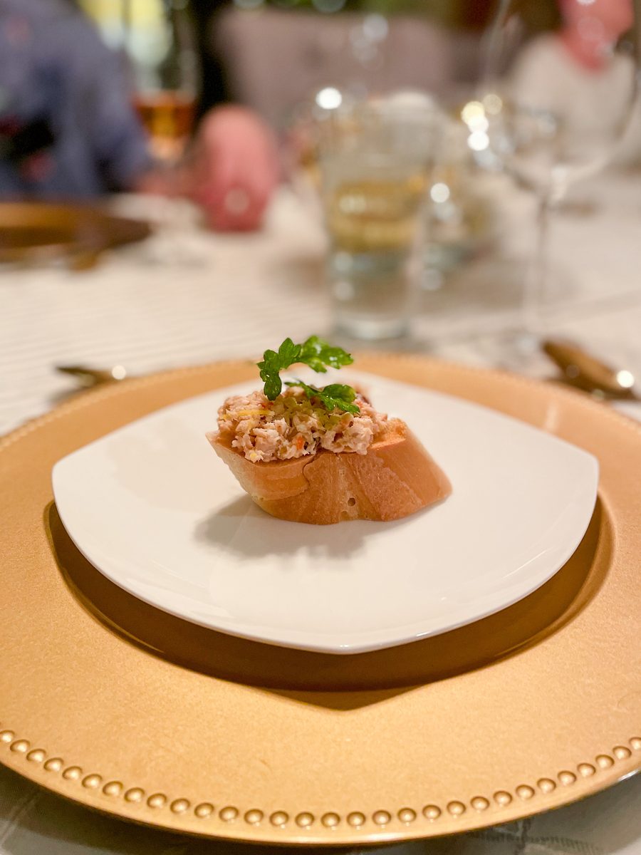 A single baguette crostini topped with a creamy shredded salad and a parsley sprig on a small white square plate over a gold charg