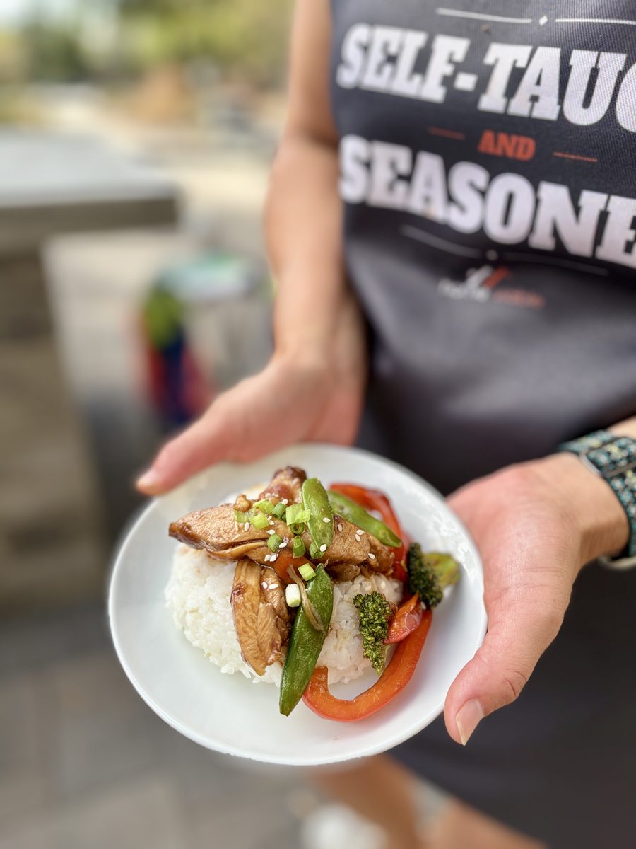 A woman in a dark graphic apron holds a small white plate of chicken stir fry with snap peas, red pepper and broccoli over white r