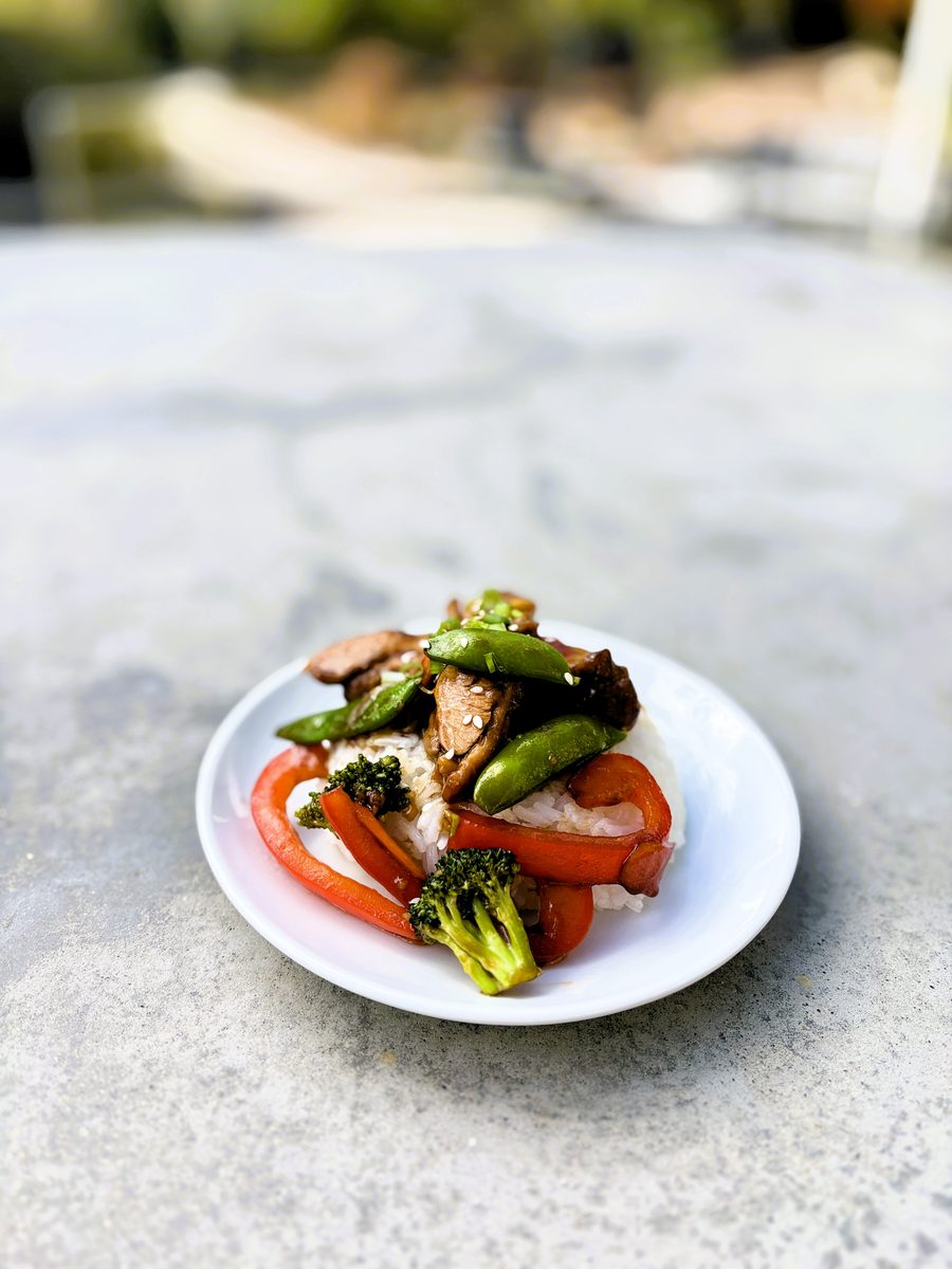 A small white plate of chicken stir fry with snap peas, red bell pepper and broccoli over white rice, sitting on a grey concrete p