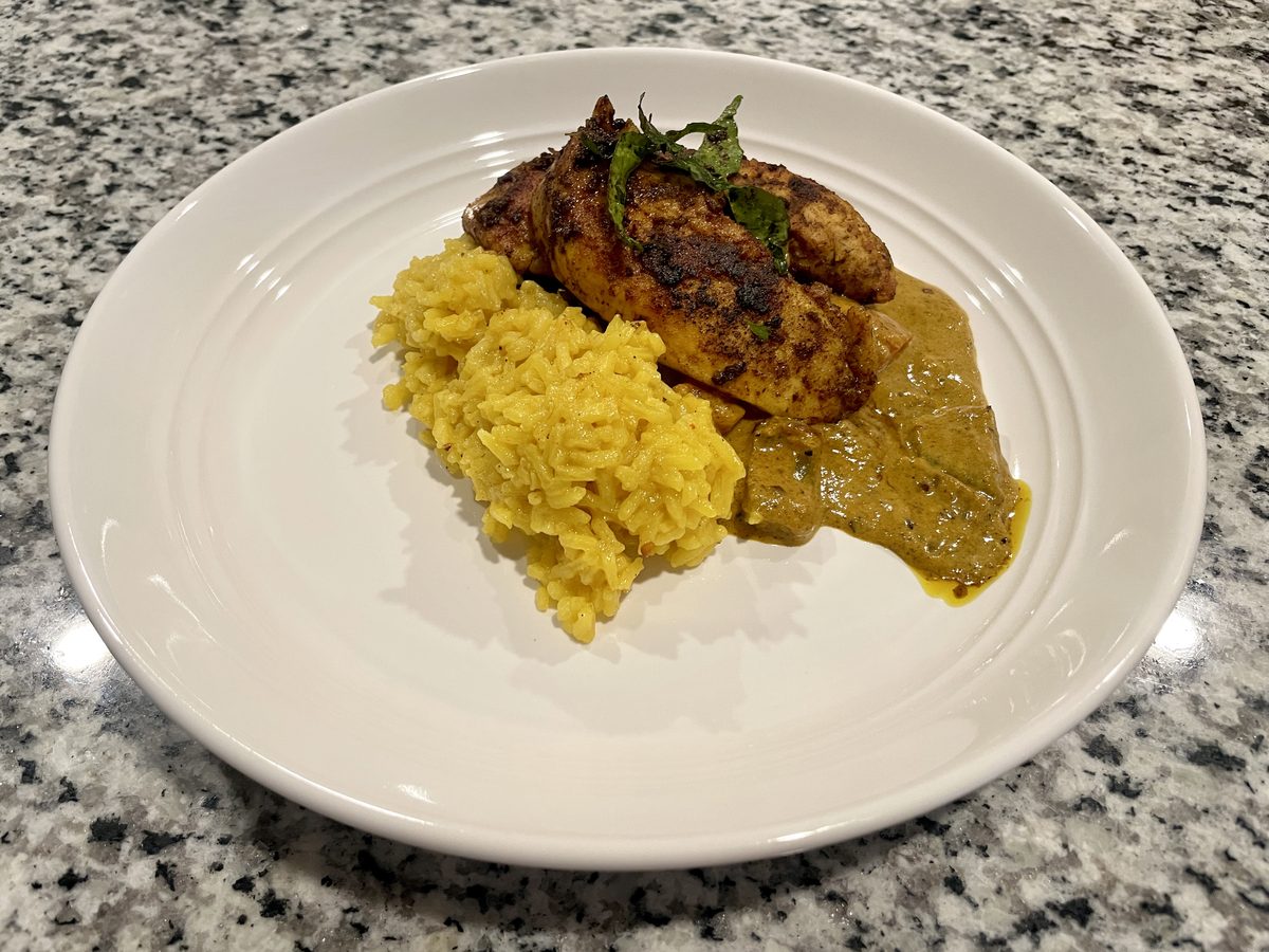 A wide white plate on a granite counter holding a charred grilled chicken breast garnished with fresh herbs, a mound of turmeric y