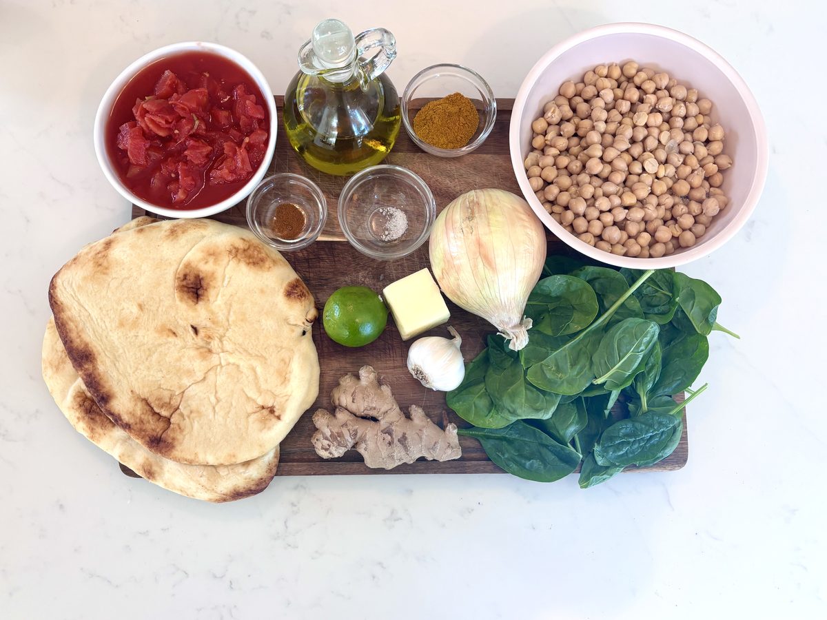 An overhead flat lay on white marble and a wooden board - a bowl of chickpeas, a bowl of crushed tomatoes, olive oil, spices in sm