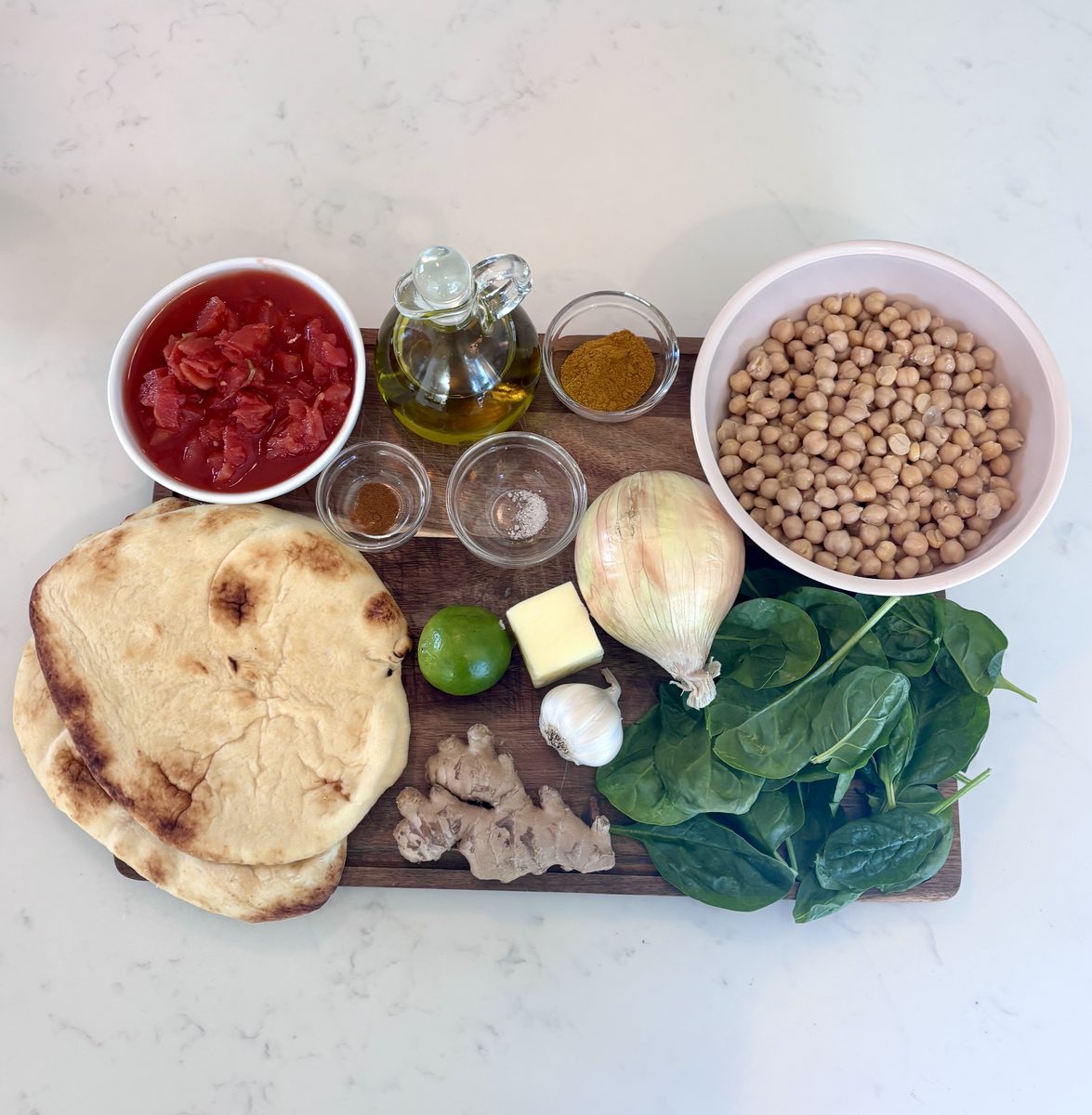 The same chickpea curry ingredient flat lay - chickpeas, crushed tomatoes, oil, spices, naan, lime, garlic, mushrooms and spinach
