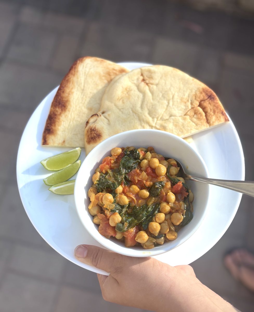 A hand holding a white plate outdoors with a white bowl of finished chickpea and spinach curry with a spoon in it, two halves of n