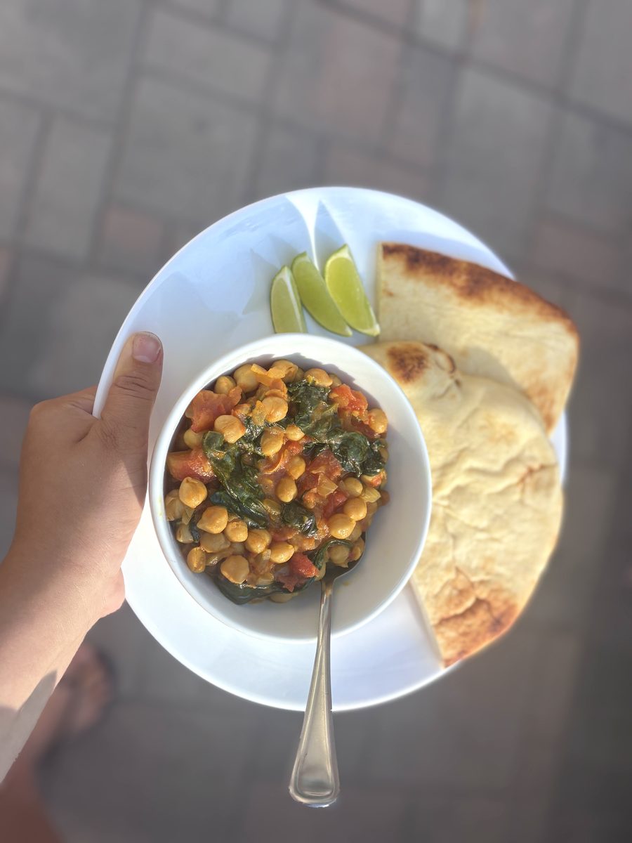 Overhead shot of chickpea and spinach curry in a white bowl on a white plate with naan and lime wedges, the plate held in a hand o